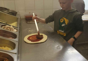 Chłopiec nakłada łyżką wazową sos pomidorowy na spód pizzy