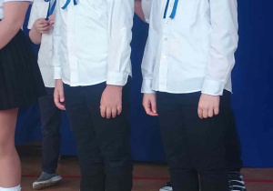 Uczniowie w strojach galowych z niebieskimi kokardami deklamują wiersz