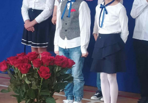 Dzieci podczas występu z okazji święta KEN na tle dekoracji i bukietu czerwonych róż