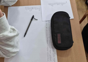 Ławka a na niej zapisana kartka z leżącym na niej długopisem, a oboki piórnik
