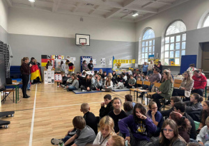 Uczniowie zgromadzeni na w sali gimnastycznej słuchający prezentacji przygotowanych przez kolegów