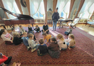 Dzieci siedzą w kręgu na dywanie w sali z fortepianem