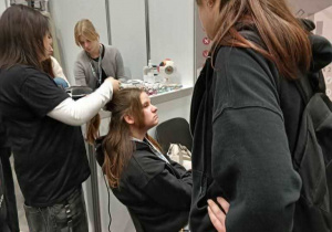 Uczennica siedzi na fotely fryzjerskim, inna dziewczyna czesze jej włosy