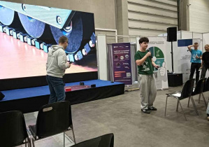 Chłopak opowiada o swoim stoisku, w tle na prezentacji na ekranie widoczna gonsienica pojazdu pancernego