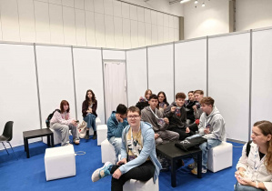 Uczniowie siedzą na białych pufach, w tle jasne moduły rozsówanej ściany, niebieska podłoga