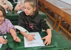 Uczniowie siedzą przy dawnych ławkach z kałamarzem i ćwiczą pisanie gęsim piórem