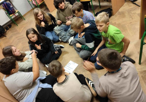 grupa uczniów siedzi na podłodze w kręgu i myśli nad zadaniem