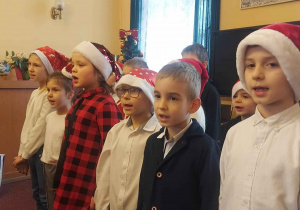 Uczniowie ustawieni w rzędzie, w mikołajowych czapkach śpiewają kolędy