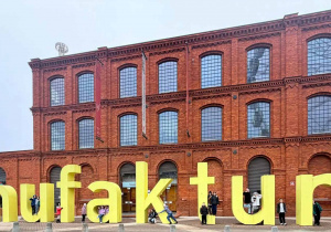 Widok ceglanego budynku a przed nim żółty napis z wielkich liter MANUFAKTURA, przy literach uczniowie