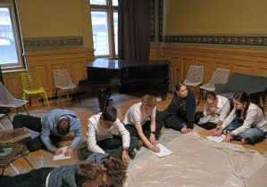 Uczniowie podczas warsztatów plastycznych - siedzą w kole na parkiecie osłoniętym folią