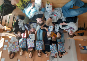 Dziewczęta stoją trzymajac w rękach torby podarunkowe, chłopcy siedzą lub leżą na parkiecie i prezentują otrzymane od dziewczyn upominki.