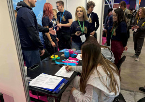 Uczennice wypełniają ankietę, w tle gospodarze stoiska