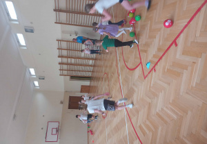 Dziecko biegnie, w tle nauczyciel w niebieskiej koszulce i inni uczestnicy na tle parkietu i drabinek