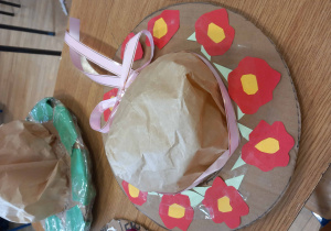 Kapelusz z kartonu z przyklejonymi czerwonymi kwiatkami z papieru, ozdobiony cienką różową wstążką