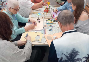 Seniorzy i wolontariusze pochyleni nad stołem tworzą świąteczne prace plastyczne