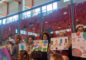 Uczniowie na widowni, w rękach trzymają plakaty ekologiczne, wszyscy ozdobieni kapeluszami wykonanymi z materiałow z recyklingu.