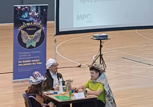 Trzy osoby z naszej drużyny siedzą przy stoliku na parkiecie w hali MOSiR, z tyłu ekran multimedialny z wyświetloną nazwą wydarzenia.