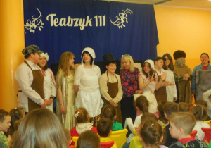 Uczniowie w kostiumach scenicznych, za nimi niebieskie tło z napisem Teatrzyk 111