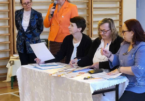 Obrady komisji konkursowej, stół przykryty tkaniną, trzy osoby siedzą, dwie panie stoją