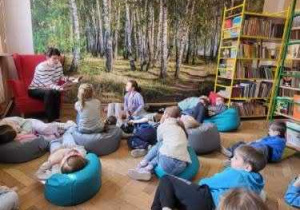 W sali bibliotecznej, uczniowie słuchają czytanej przez nauczycielkę lektury siedząc lub leżąc wygodnie na niebieskich pufach