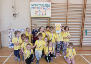 Żółci prezentują swoje nagrody na tle plakatu z napisem "Olimpiada Przedszkoli"