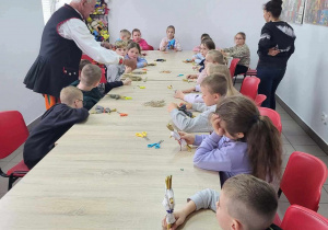 Zajęcia z łowickiej wycinanki- dzieci siedzą przy długim, jasnymstole- pan w stroju ludowym rozdaje im pomoce