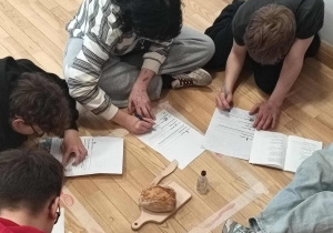 Uczniowie pochyleni nad kartkami z pracą przed nimi w oznaczonym jak w kryminale polu leży drewniana deska do krojenia na niej bułka a obok mały nożyk