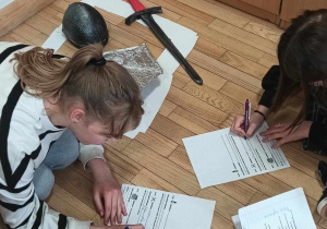 Uczennice wpisują odkryte treści do swoich kart pracy, w tle dowody zbrodni - miecz