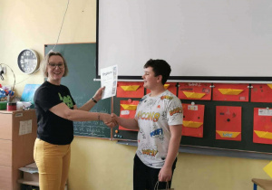 Pani Magdalena gratuluje zwycięzcy konkursu, w tle biały ekran a pod nim dekoracja w postaci czerwonych kart z Żółtymi łódeczkami z origami, nawiązanie do herbu miasta Łodzi