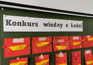 Dekoracja- napis na białym tle Konkurs wiedzy o Łodzi, pod spodem żółte łódeczki z origami na czerwonym polu - nawiązanie do herbu miasta
