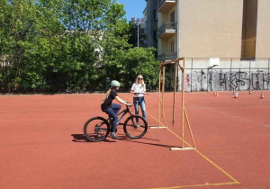 Na tle brązowej nawierzchni boiska rowerzytka pokonuje przeszkodę podczas egzaminu praktycznego na kartę rowerową