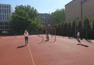 Na tle brązowej nawierzchni boiska rowerzytka pokonuje przeszkodę podczas egzaminu praktycznego na kartę rowerową, a nauczycielka/egzaminatorka idzie obok.