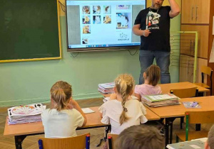 Wolontariusz na tle ekranu multmedialnego z prezentacją, na pierwszym planie uczniowie siedzący w ławkach.