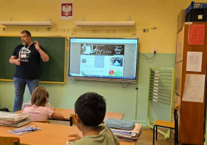 Wolontariusz na tle ekranu multmedialnego z prezentacją, na pierwszym planie uczniowie siedzący w ławkach.