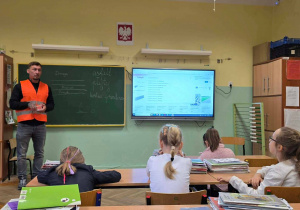Mężczyzna w odblaskowej, pomarańczowej kamizelce stoi przy zielonej tablicy, uczniowie siedzący w ławkach uważnie go słuchają.