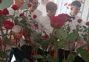 Uczniowie trzymają czerwone róże połączone w okrąg bordową wstążką