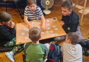 Dzieci siedzą przy kwadratowym stoliku z jasnego drewna i mają przed sobą fiszki potrzebne do zajęć
