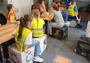 Dzieci w odblaskowych kamizelkach siedzą na pufach i słuchają